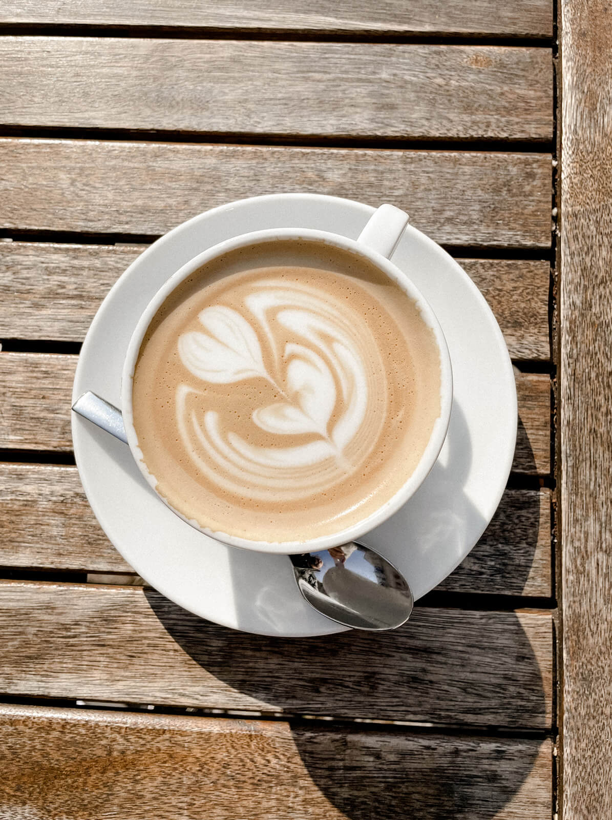 Cappuccino mit einer Blume aus Milchschaum auf einem Holztisch im Sonnenschein.